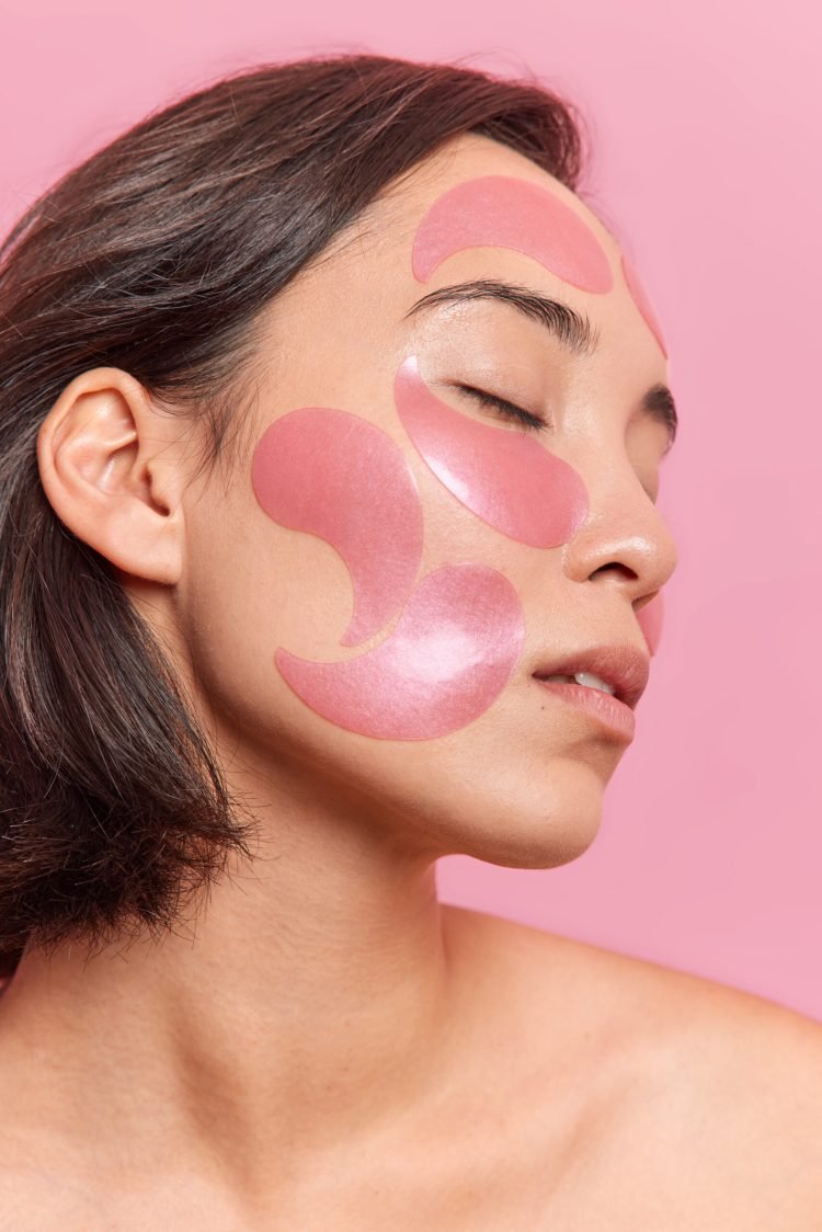 Profile shot of brunette Asian woman keeps eyes closed applies hydrogel patches on face to refresh skin has healthy well cared skin dark short hair isolated over pink backgroud. Beauty concept